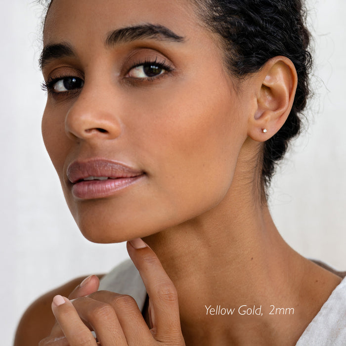 Close-up of a woman wearing 14k Yellow Gold Diamond Stud Earrings 2mm - Aria with natural makeup on a white background