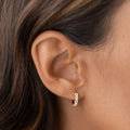 Close-up of an ear wearing April Birthstone Huggie Earrings - White Topaz with gold vermeil setting on neutral background