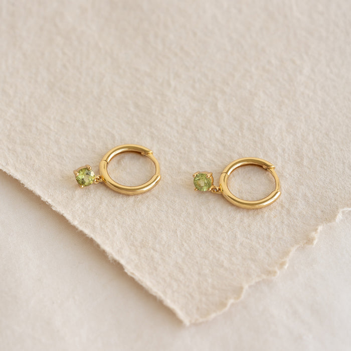 Close-up of gold vermeil August Birthstone Drop Huggie Earrings - Peridot with green gemstones on textured beige paper