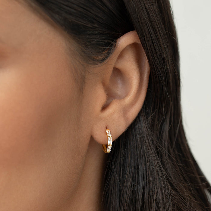 Close-up of a woman’s ear wearing a small Gold Vermeil earring set with baguette-cut gemstone, shown against a neutral background with dark hair tucked behind the ear.