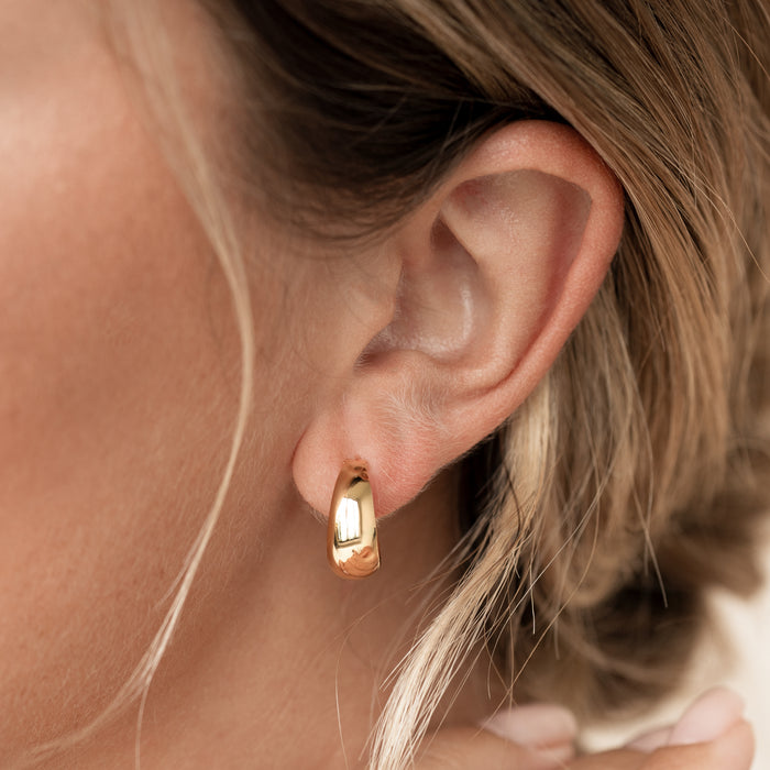 A close-up profile shot focusing on a person's left ear, showcasing a chunky, high-polish gold huggie hoop earring on the lobe.