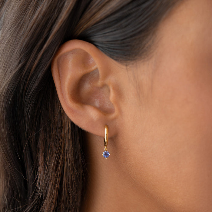 Close-up of a person’s ear wearing a small Gold hoop earring with a dangling round light blue gemstone, shown against smooth skin and dark brown hair.