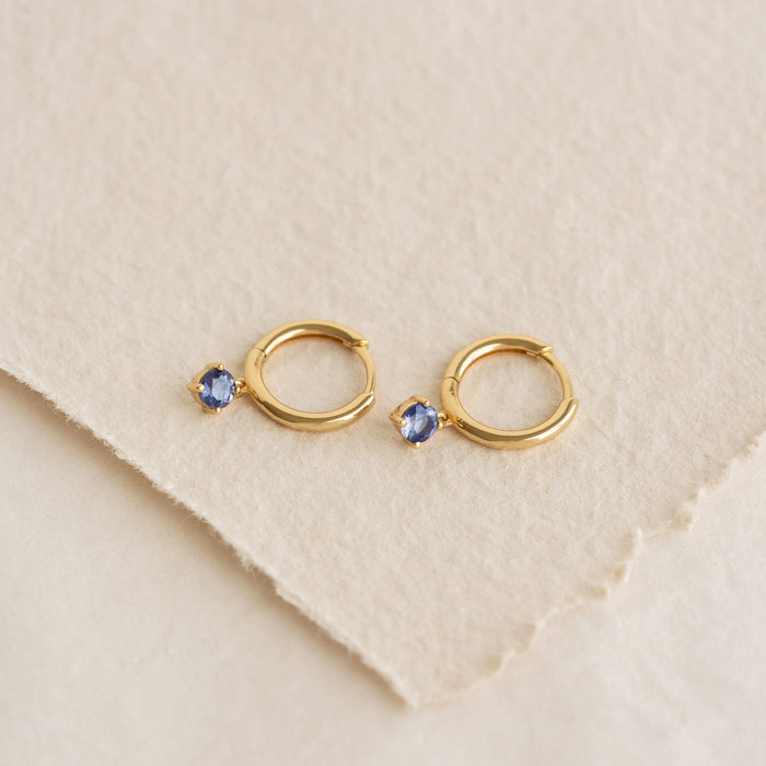 Pair of small Gold hoop earrings with dangling oval light blue gemstones, placed on a soft, textured off-white fabric surface.