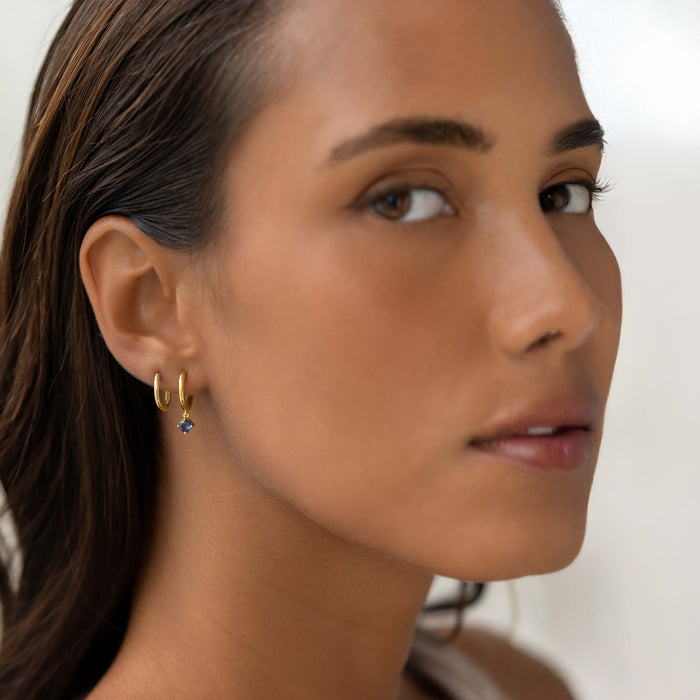 Close-up of a woman wearing December Birthstone Drop Huggie Earrings - Cordierite with gold vermeil setting on a neutral background