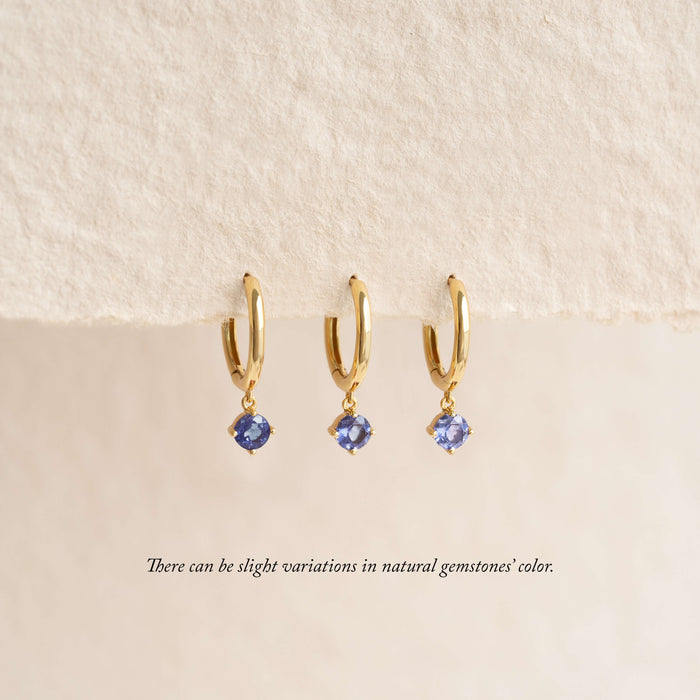 Three Gold hoop earrings with small dangling light blue gemstones are displayed hanging from a soft, textured fabric background, with a note below indicating that natural gemstone colors may vary.