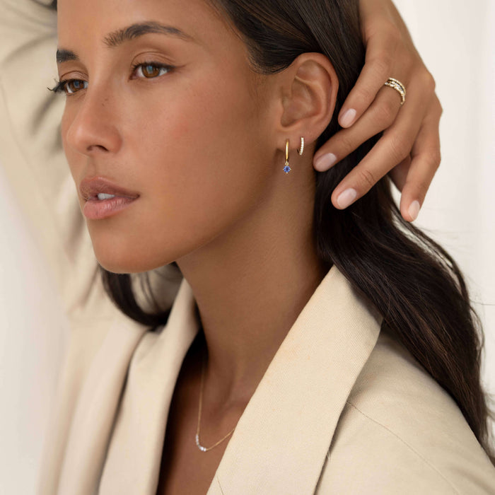Close-up of a woman wearing two gold earrings—a small hoop with a dangling light blue gemstone and a textured huggie—while gently touching her ear, paired with a delicate necklace and neutral-toned outfit.