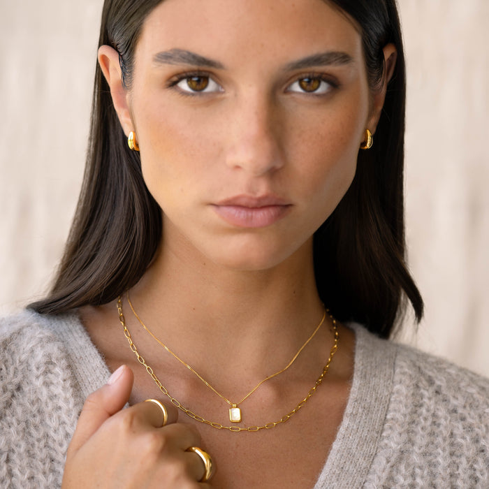 Close-up of a woman wearing Dome Huggie Earrings - Caja in gold vermeil, paired with gold necklaces and rings against a neutral background