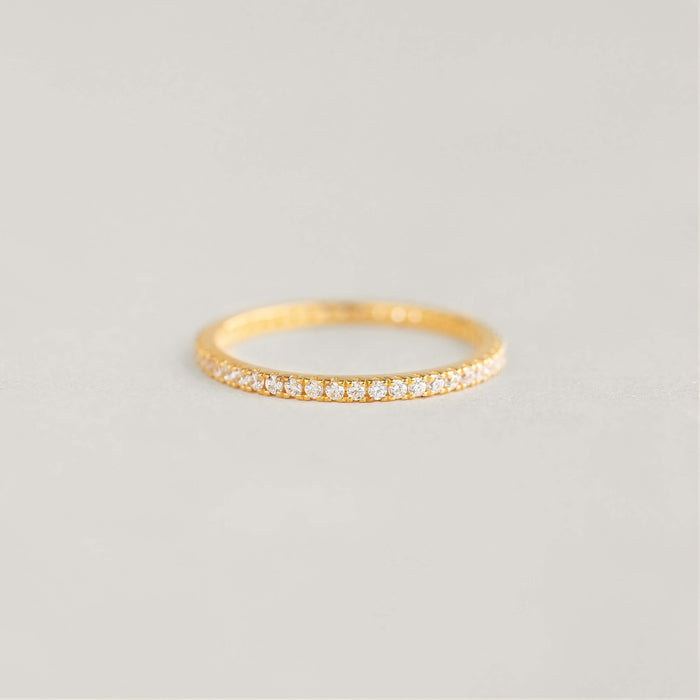 A delicate Gold band ring with small diamonds encircling it is placed on a soft, neutral background, exuding elegance and simplicity.