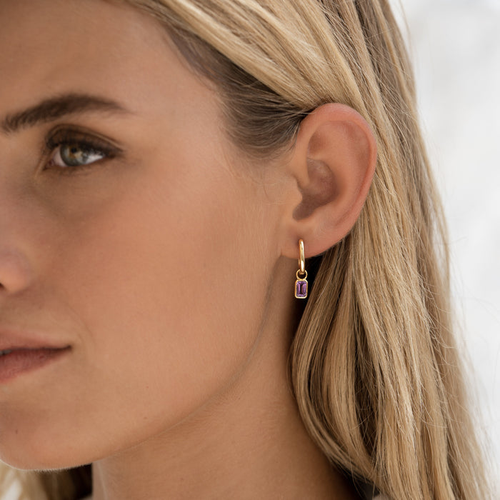 Close-up of woman wearing Huggie Earrings with Baguette Charms Amethyst - Amelia in gold vermeil on neutral background