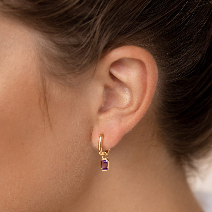 Close-up of a woman wearing Huggie Earrings with Baguette Charms Amethyst - Amelia on a neutral background