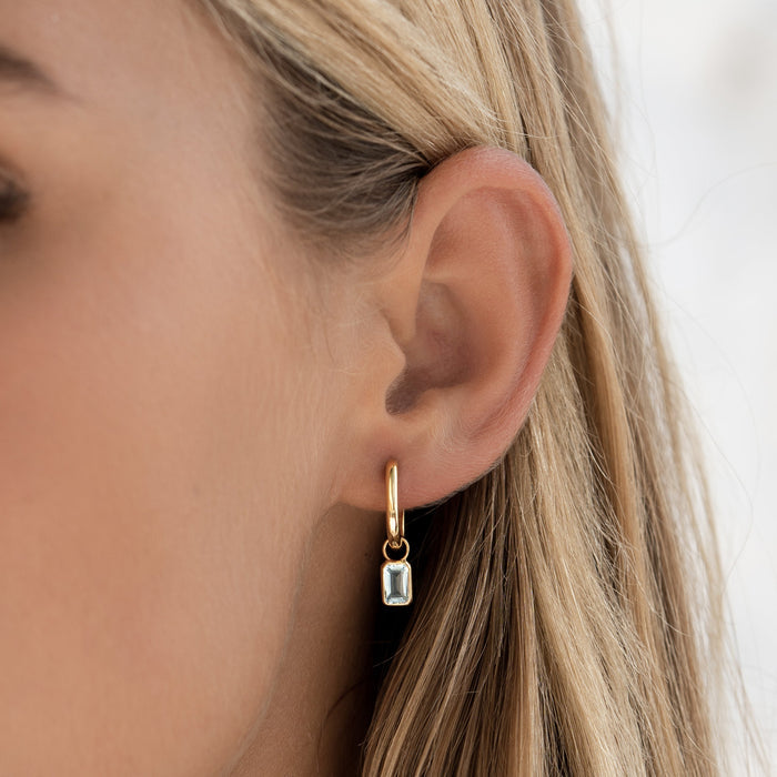 Close-up of an ear wearing Huggie Earrings with Baguette Charms Sky Blue Topaz - Amelia in gold vermeil