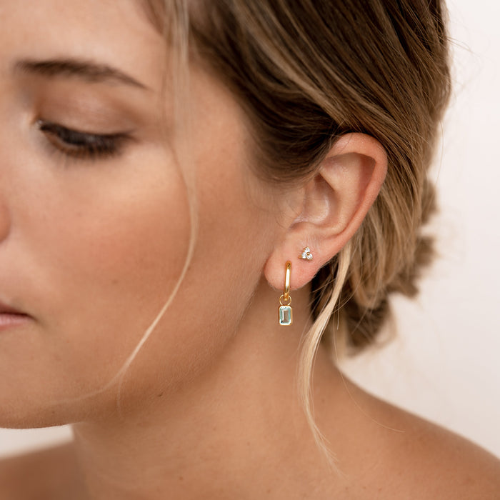 Close-up of a woman wearing Huggie Earrings with Baguette Charms Sky Blue Topaz - Amelia in gold vermeil on a neutral background