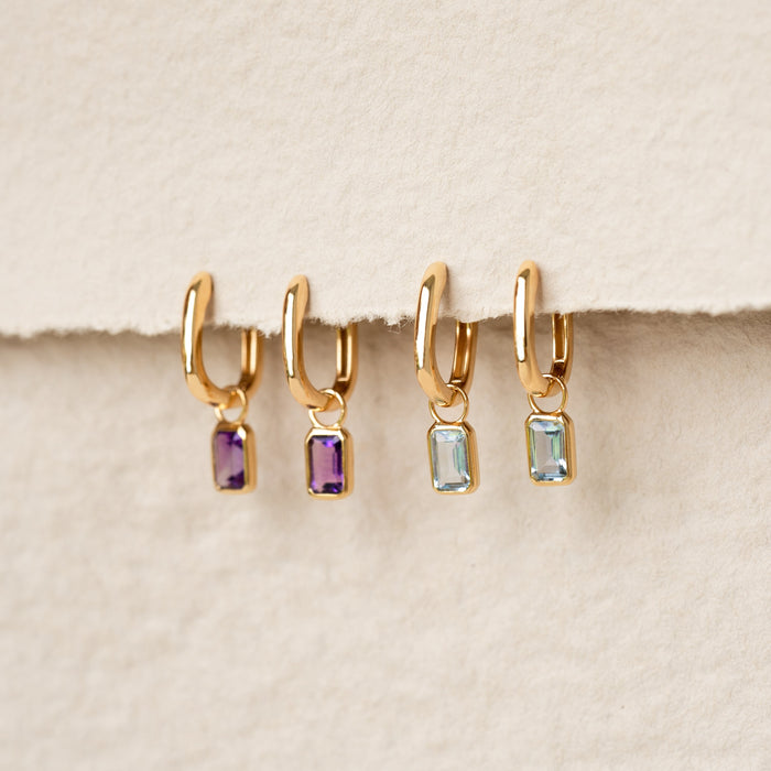 Four gold vermeil Huggie Earrings with Baguette Charms Sky Blue Topaz - Amelia and purple stones on a beige background