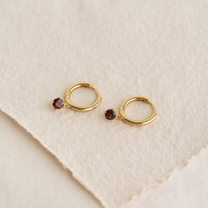 Close-up of January Birthstone Drop Huggie Earrings - Garnet with gold vermeil setting on a textured beige background