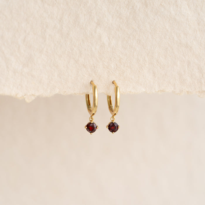 Close-up of January Birthstone Drop Huggie Earrings - Garnet with gold vermeil setting on a neutral textured background