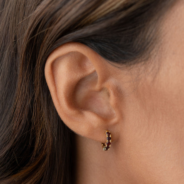 Close-up of a woman's ear wearing January Birthstone Huggie Earrings - Garnet with gold vermeil setting on neutral background