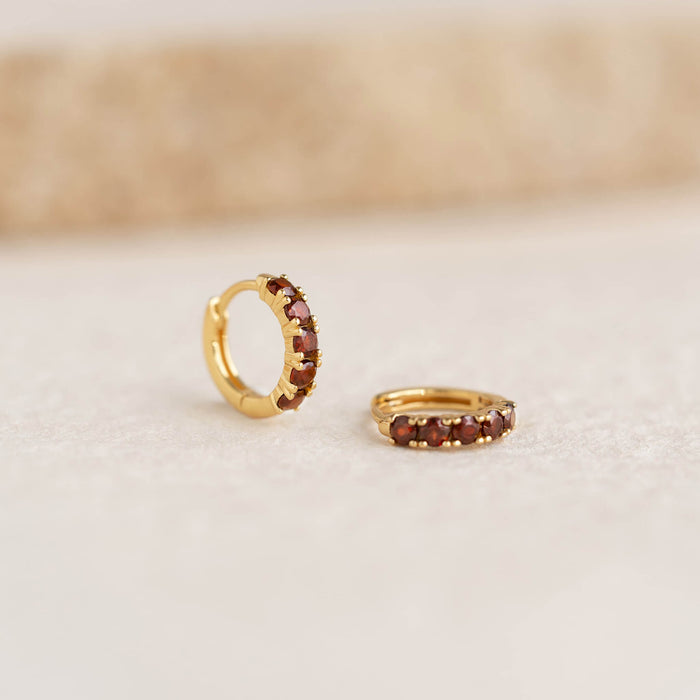 Close-up of January Birthstone Huggie Earrings - Garnet with gold vermeil and deep red gemstones on a light surface