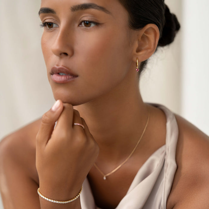 Woman wearing July Birthstone Drop Huggie Earrings - Pink Tourmaline with gold necklace and bracelet on neutral background