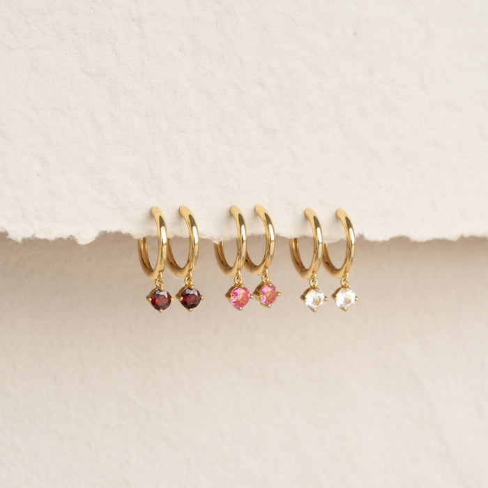 Three pairs of July Birthstone Drop Huggie Earrings - Pink Tourmaline and other gemstones with gold vermeil hoops on beige background
