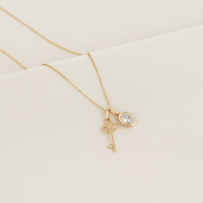 Gold necklace featuring a key-shaped charm with a small diamond accent and a oval diamond pendant, displayed on a white textured surface.