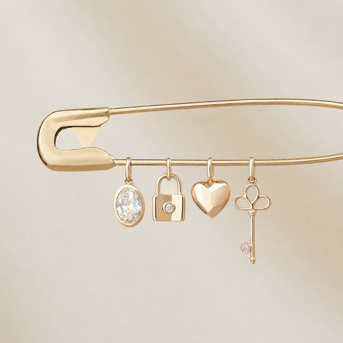 Yellow Gold safety pin featuring four hanging charms: an oval diamond charm, a padlock with a round diamond, a heart-shaped charm, and a key charm with a small diamond accent.