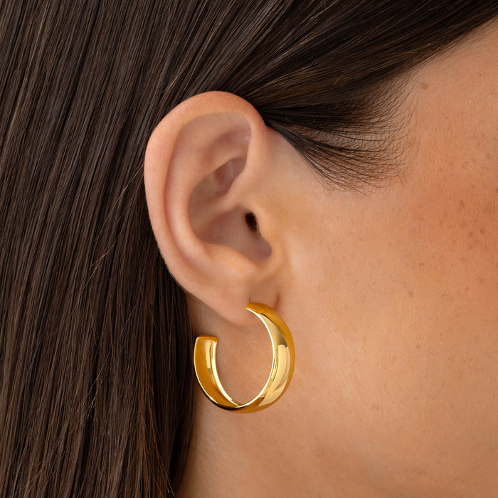 Close-up of a woman’s ear wearing Large Bold Hoop Earrings - Mina in polished gold vermeil on a neutral background