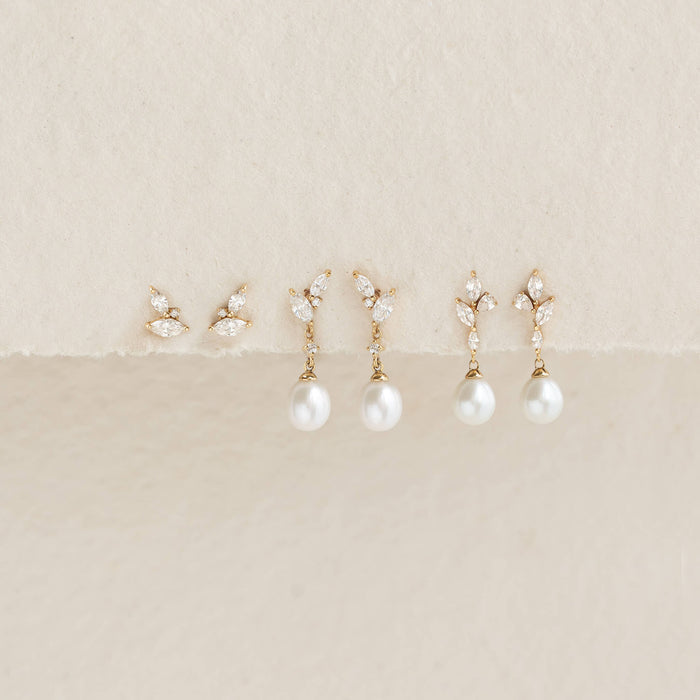 A collection of elegant earrings is displayed on a soft beige background. The earrings feature Gold leaves with sparkling stones, and some have dangling pearls.