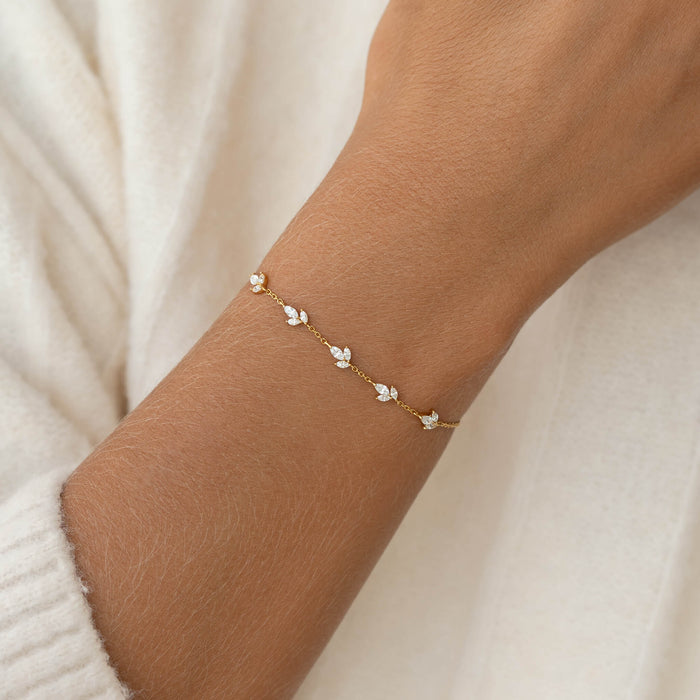 A delicate Gold bracelet with small white gemstones is elegantly worn on a wrist, framed by soft beige fabric, conveying a refined and minimalist style.