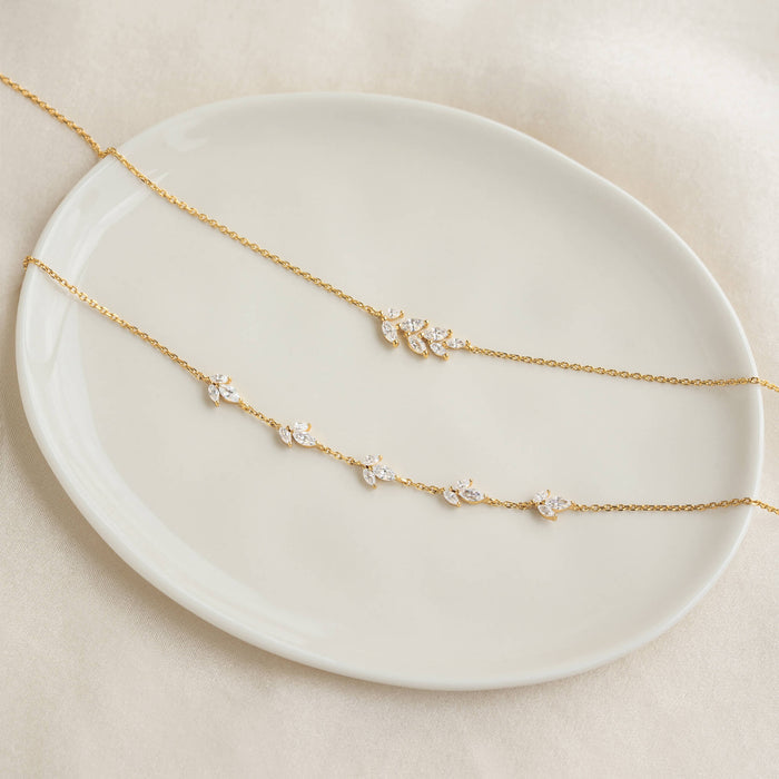 Two delicate Gold necklaces with leaf-like crystal accents are elegantly displayed on a smooth, white ceramic plate. The setting is soft and minimalistic.