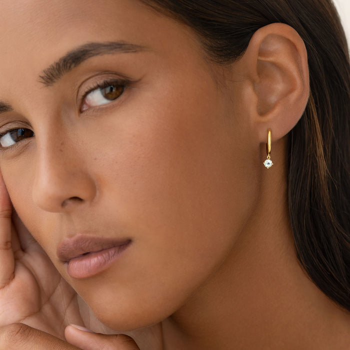 Close-up of a woman wearing March Birthstone Drop Huggie Earrings - Aquamarine with gold vermeil setting and pale blue gemstone
