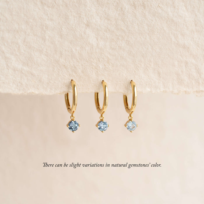 Three gold hoop earrings with small dangling light blue gemstones are displayed hanging from a soft, textured fabric background, with a note below indicating that natural gemstone colors may vary.
