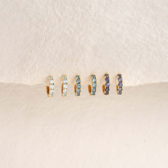 Three pairs of March Birthstone Huggie Earrings - Blue Topaz featuring gold vermeil and blue gemstones on a textured background