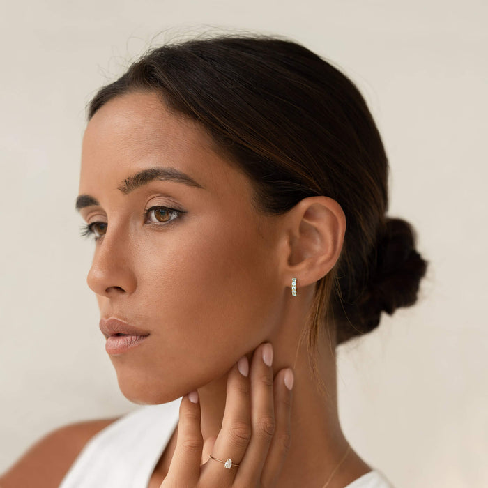 Young woman wearing March Birthstone Huggie Earrings - Blue Topaz with hair in a low bun against neutral background