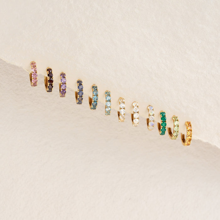 Row of colorful huggie earrings with various gemstones including March Birthstone Huggie Earrings - Blue Topaz on a light textured background