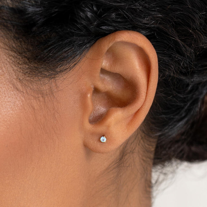 Close-up of an ear wearing March Birthstone Stud Earrings 14k Gold - Blue Topaz with a soft neutral background