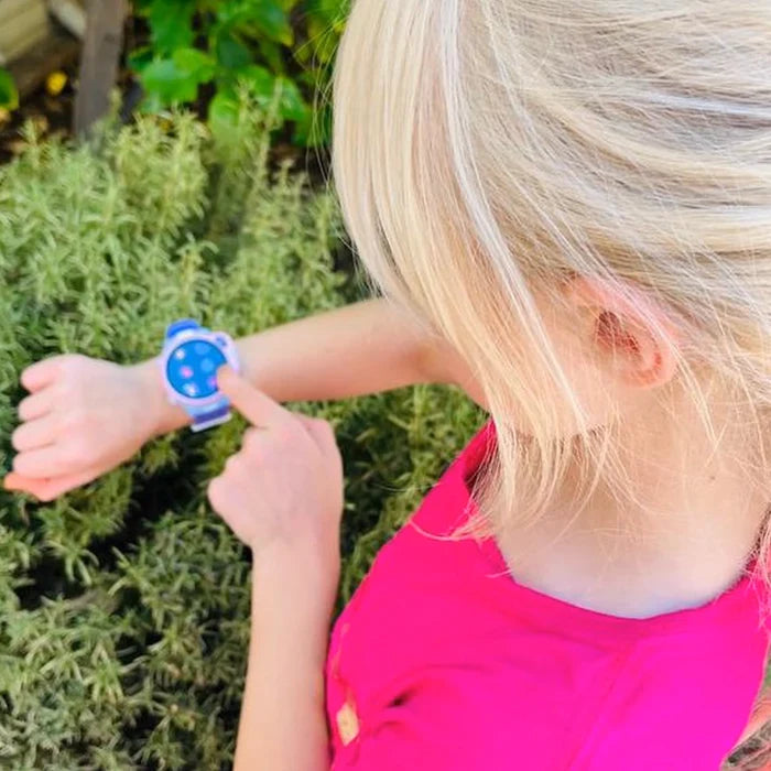 myFirst Fone R2 kids smartwatch worn by a young girl in a pink top, pointing at the watch outdoors in a garden setting.