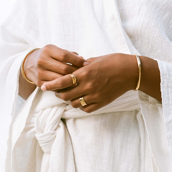 Close-up of hands wearing gold bangles and the Organic Wavy Ring - Bea with a white textured robe background