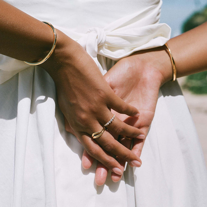 Hands crossed at the waist wearing the Organic Wavy Ring - Bea and gold bangles over a white dress in natural light