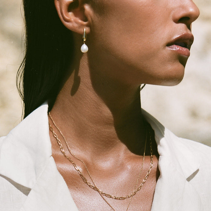 Close-up of a woman wearing Pearl Drop Hoop Earrings - Maeve in gold vermeil with layered gold necklaces and white blouse