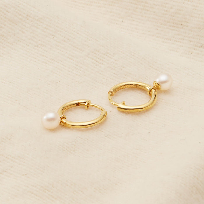 Gold hoop earrings with small white pearls on a soft beige fabric. The elegant and minimalist design adds a touch of luxury and sophistication.