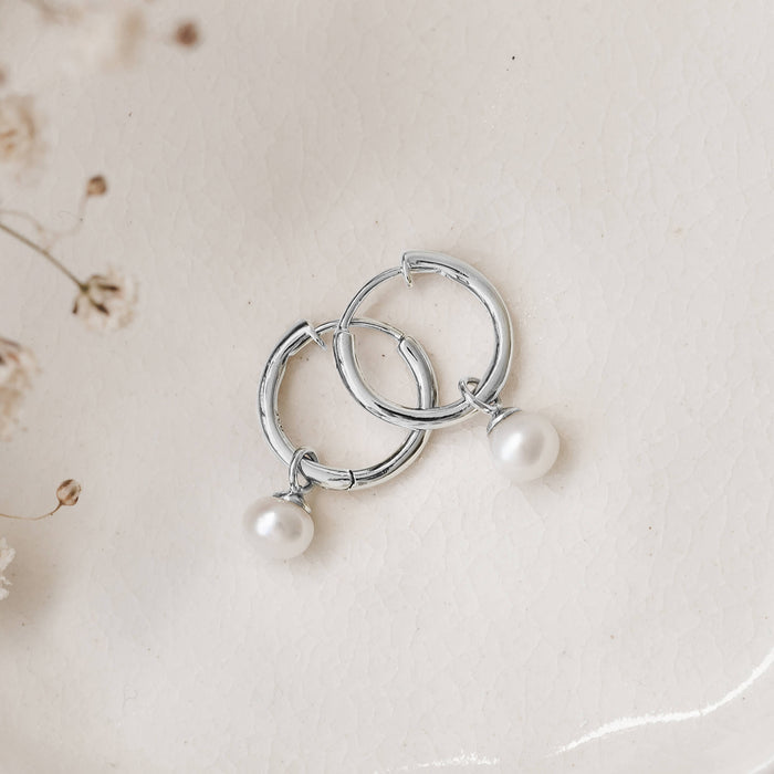 Silver hoop earrings with dangling pearls lie on a textured, light cream surface. Delicate dried flowers are visible in the top left corner.