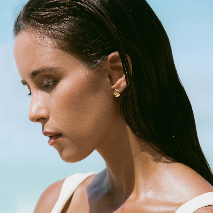 Close-up of a woman wearing Scalloped Huggie Earrings - Tulisa in gold vermeil against a soft blue sky background