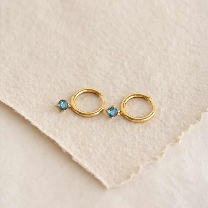 Close-up of September Birthstone Drop Huggie Earrings - London Blue Topaz with gold vermeil setting on textured paper background