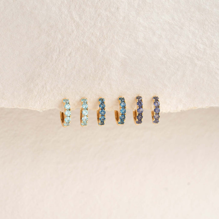 September Birthstone Huggie Earrings - London Blue Topaz in gold vermeil with varying blue gemstones on a cream background