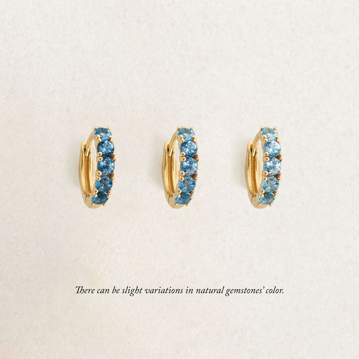Three gold hoop earrings set with blue gemstones are displayed upright in a row on a light background. Text below notes slight color variations in natural gemstones.