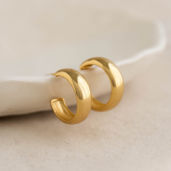 Close-up of Small Bold Hoop Earrings - Ebba in gold vermeil displayed on a neutral textured surface