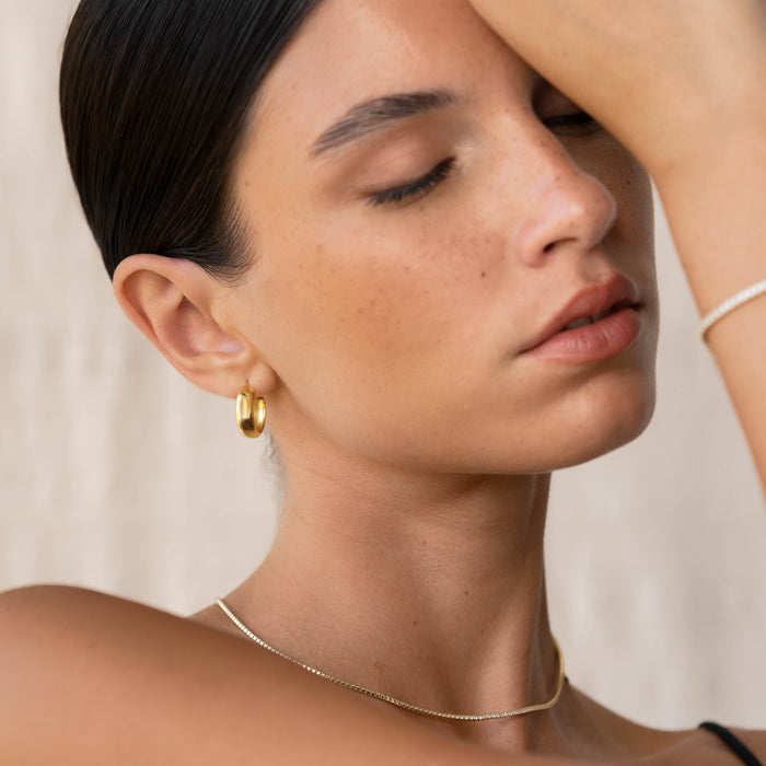 Close-up of a woman wearing Small Bold Hoop Earrings - Ebba in gold vermeil with a delicate gold necklace against a neutral background