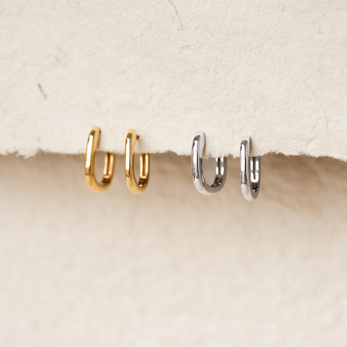 Square Huggie Earrings Silver - Amelia with a playful design displayed alongside gold earrings on a neutral textured background