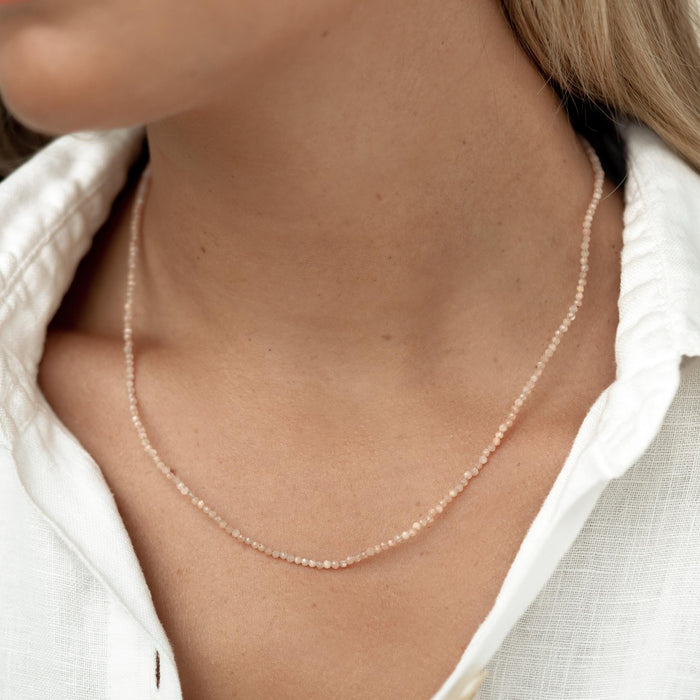 Close-up of a woman wearing a delicate beaded Sunstone Necklace with soft peach tones and a white linen top