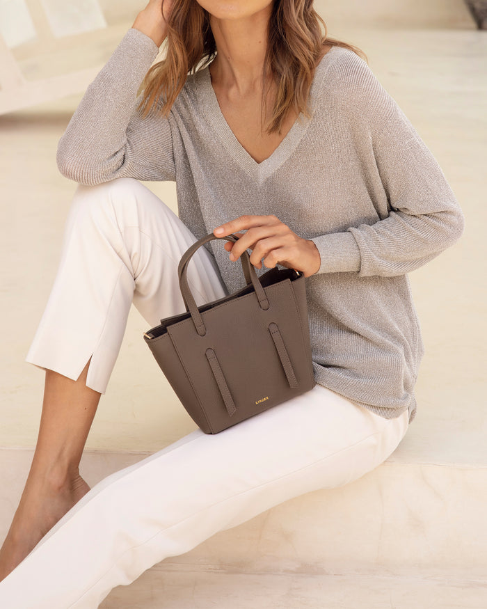 Woman seated wearing light gray sweater and white pants holding The Lana Bag - Blue with top handles against neutral background